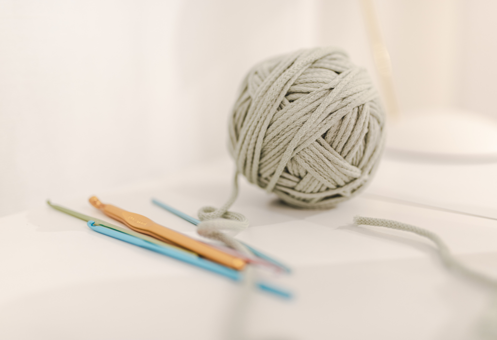 A ball of light gray cotton yarn for crochet sits beside three crochet hooks on a white surface.